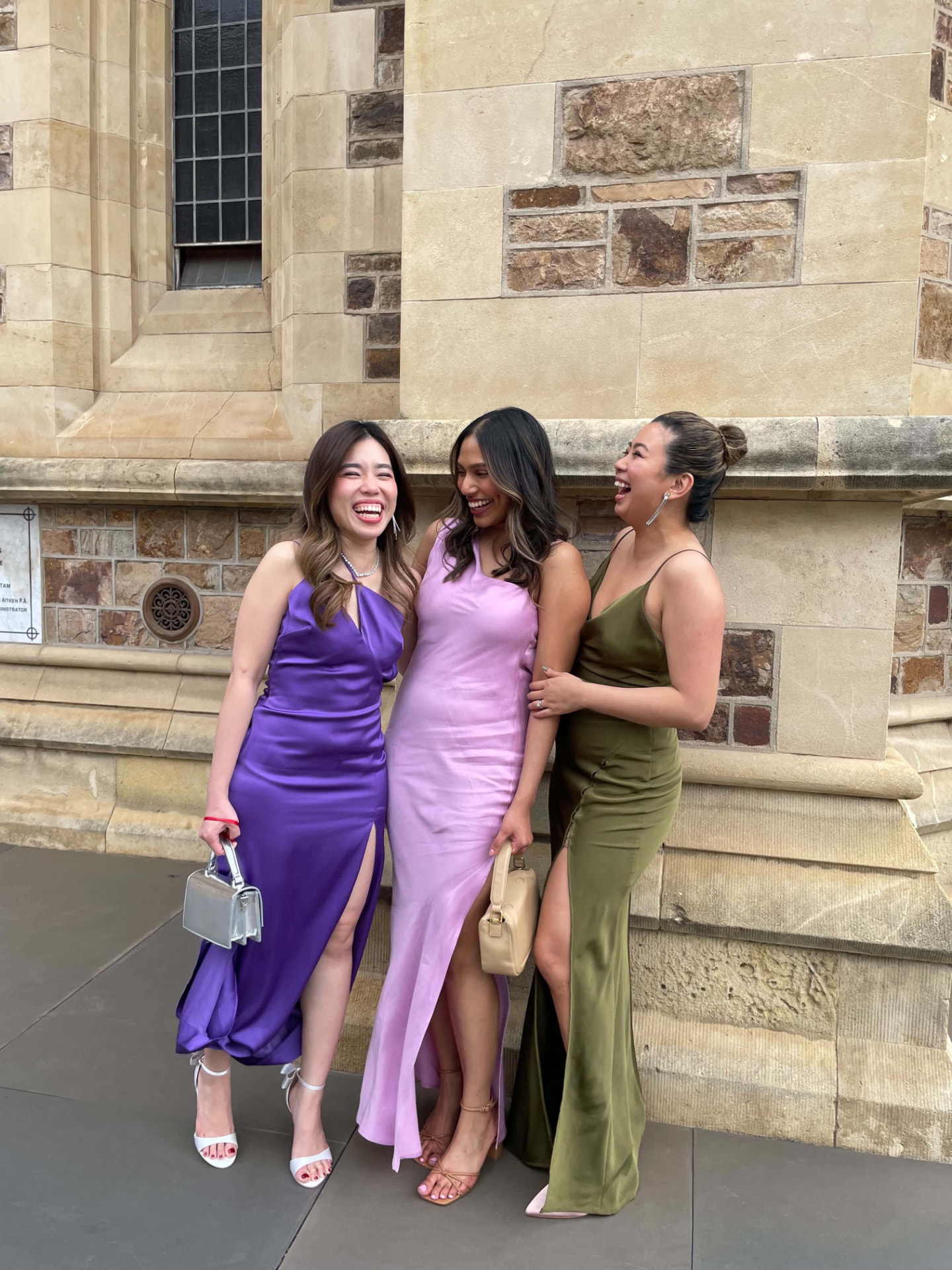 Three friends in satin gowns laughing together outside a stone cathedral